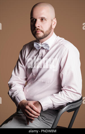 Vue côté profil portrait of handsome man chauve barbu en chemise rose et arc blanc, assis sur une chaise et à l'écart avec sérieux attentionné f Banque D'Images