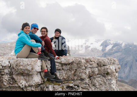 Quatre jeunes randonneurs masculins et féminins assis sur un pic de montagne rocheuse dans les Dolomites et à la recherche à la vue incroyable avec Langkofel et sommets Marmolada Banque D'Images