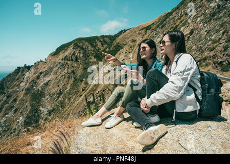 Deux femmes touristes trouver quelque chose par le téléphone et une fille est pointing à venir tout en restant assis sur la falaise. Banque D'Images