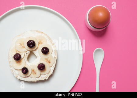 La moitié de bagel sur plaque blanche avec fromage à la crème, les bleuets, et des tranches de banane avec un oeuf dur et cuillère blanc sur fond rose Banque D'Images