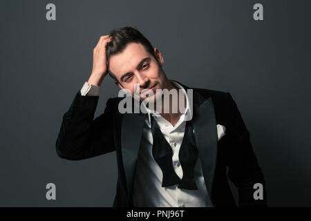 Portrait noir et blanc d'un jeune homme séduisant en costume, souriant et regardant la caméra, noeud papillon et boutons de chemise défait Banque D'Images