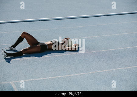 Athlète femme couchée sur la piste de course de détente après la formation. Sprinter femelle reposant après entraînement située sur la voie sur une journée ensoleillée. Banque D'Images