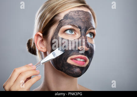 Jeune femme l'application de charbon activé, masque de visage à l'aide d'un pinceau sur fond gris Banque D'Images