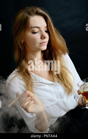 Jeune femme assise avec un cigarillo et verre de whisky à la main. Femme élégante avec haut blanc et robe noire. Banque D'Images