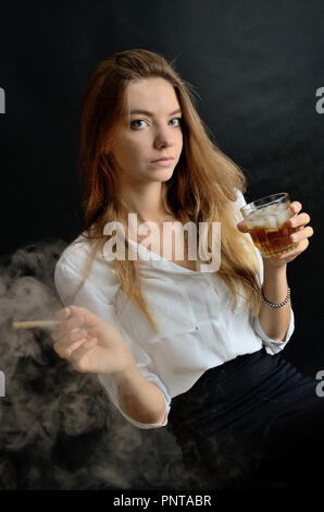 Jeune femme assise avec un cigarillo et verre de whisky à la main. Femme élégante avec haut blanc et robe noire. Banque D'Images