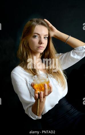 Jeune femme assise avec un cigarillo et verre de whisky à la main. Femme élégante avec haut blanc et robe noire. Banque D'Images