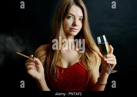 Jeune femme assise avec un cigarillo et verre de vin blanc dans sa main. Femme en robe de soirée rouge. Banque D'Images