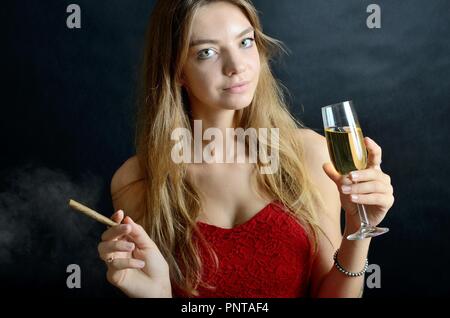 Jeune femme assise avec un cigarillo et verre de vin blanc dans sa main. Femme en robe de soirée rouge. Banque D'Images