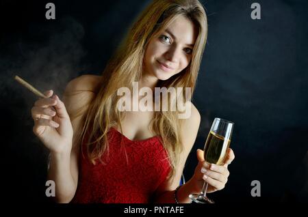 Jeune femme assise avec un cigarillo et verre de vin blanc dans sa main. Femme en robe de soirée rouge. Banque D'Images