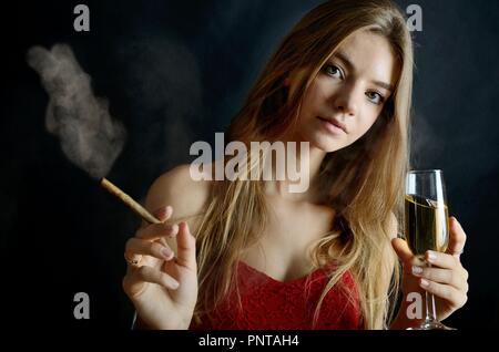 Jeune femme assise avec un cigarillo et verre de vin blanc dans sa main. Femme en robe de soirée rouge. Banque D'Images