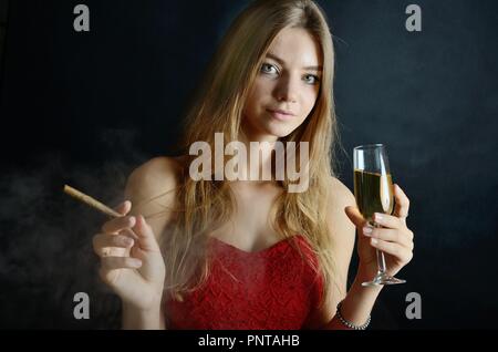 Jeune femme assise avec un cigarillo et verre de vin blanc dans sa main. Femme en robe de soirée rouge. Banque D'Images