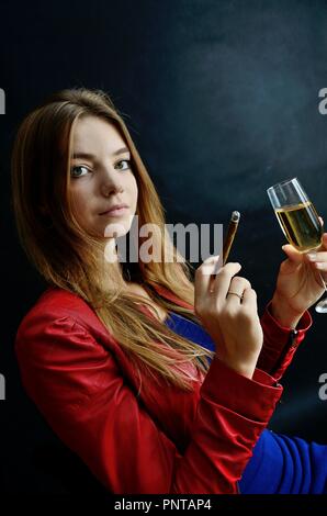 Jeune femme assise avec un cigarillo et verre de vin blanc dans sa main. Woman in red jacket. Banque D'Images