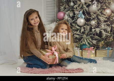 Deux petites soeurs sont en attente de Noël. Les filles sont assis à l'arbre de Noël, smiling, cadeaux. Ils sont habillés en jeans et pulls. Banque D'Images