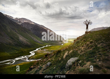 Capture d'Epic Homme tenant son vtt sur la colline en silhouette avec la rivière dans la vallée de montagne l'arrière-plan. Banque D'Images