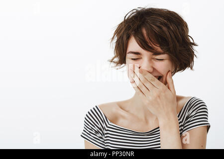 Vous arrêter de me faire rire. Portrait of happy joyeux désordre fille mignonne avec des cheveux courts et joli imprimé rayé top, rire avec les yeux fermés, riant et couvrant la bouche avec palm Banque D'Images