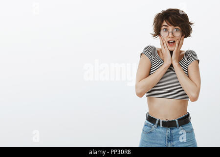 Portrait de femme européenne heureux surpris avec les cheveux bruns courts à lunettes rondes et vêtement élégant, haletant de l'intérêt, holding palms près de l'embouchure, d'être impressionné sur fond gris Banque D'Images