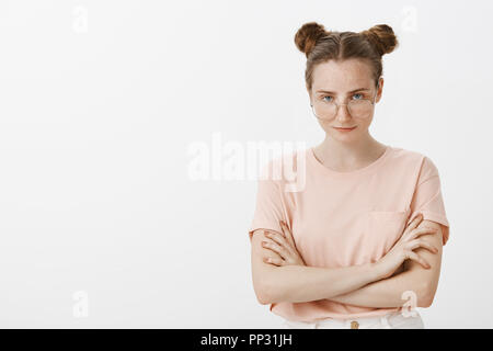 Femelle européenne arrogante Smart blogger dans l'élégant T-shirt rose avec des brioches à la coiffure, à partir de sous front lors du port de lunettes à la mode, debout avec les mains croisées sur la poitrine, sentiment toujours raison Banque D'Images