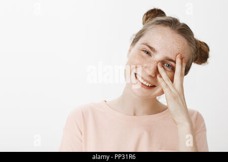 Gros plan d'étudiante insouciante heureux en t-shirt rose avec brioches hairstyle, taches de rousseur et mignon yeux bleus, tête inclinable et peeking through fingers à huis clos avec un large sourire magnifique. Banque D'Images