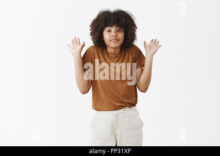 Je n'ai pas de s'en occuper. Portrait de calme à l'écart et désintéressé african american woman avec les cheveux bouclés en haussant avec palmiers ayant soulevé aucune idée de sujet sur fond gris Banque D'Images