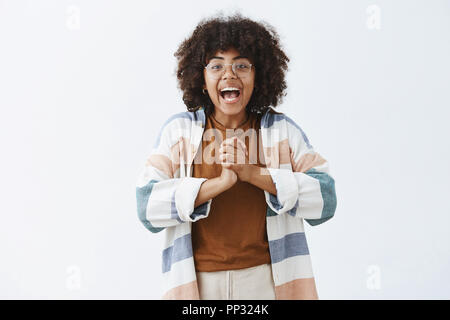 Active enthousiaste et insouciante jeune fille optimiste à la peau sombre dans les verres et les mains jointes tenue élégante près de poitrine et le sourire d'être excité avec une excellente nouvelle pour lui dire de reste de personnes Banque D'Images