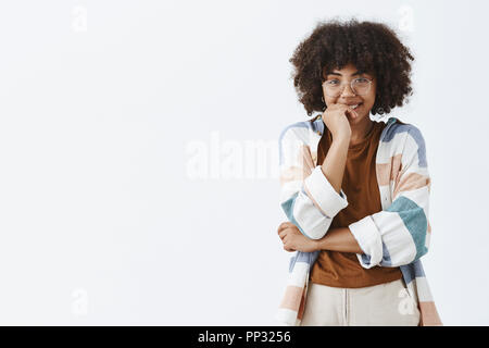 Tourné à l'intérieur de silly et flirty joli modèle féminin à peau foncée avec curly hairstyle dans verres transparents et tendance avec un large sourire tenue couvrant la bouche avec de l'expression timide palm Banque D'Images