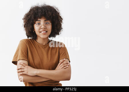 Jusqu'à la taille de balle et timide à peau sombre idiote adolescente en verres transparents et tendance brown t-shirt sensations de froid traversant les mains sur la poitrine et souriant avec expression aimable et poli Banque D'Images