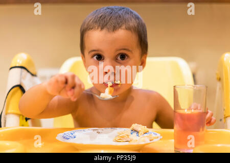 Le garçon de 2 ans mange de la viande. Table enfant. Le concept de l'indépendance de l'enfant. Mignon bébé garçon avec cuillère est yogourt. l'enfant sourit. funny k Banque D'Images