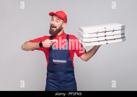 Les jeunes professionnels delivery man with beard en uniforme bleu et t-shirt rouge, debout et face à la pile de boîtes à pizza en carton gris sur ba Banque D'Images