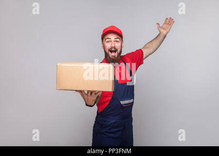 Les jeunes heureux homme livraison logistique avec barbe en uniforme bleu et t-shirt rouge, permanent holding cardboard box et souriant à pleines dents sur fond gris. Dans Banque D'Images