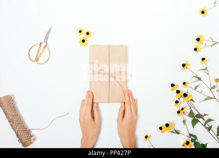 Vue de dessus les mains tiennent un présent près de fleurs jaune vif, des ciseaux d'or et de la ficelle sur le tableau blanc. Télévision Floral composition laïcs femme se donnent la main Banque D'Images