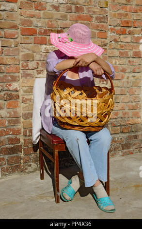 Close-up portrait of mature woman in chapeau à large qui est assis sur la chaise baissant la tête plus grand panier en osier sur ses genoux en Banque D'Images