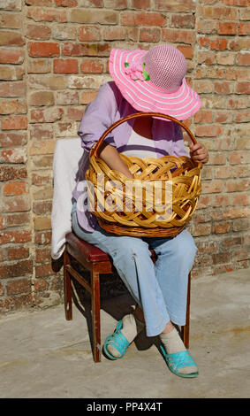 Close-up portrait of mature woman in chapeau à large qui est assis sur la chaise baissant la tête plus grand panier en osier sur ses genoux en Banque D'Images