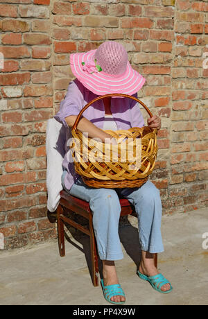 Close-up portrait of mature woman in chapeau à large qui est assis sur la chaise baissant la tête plus grand panier en osier sur ses genoux en Banque D'Images