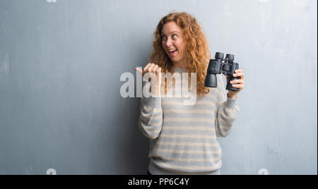 Jeune femme rousse sur mur gris grunge binoculars pointant et montrant avec le pouce jusqu'à la côte avec happy face smiling Banque D'Images