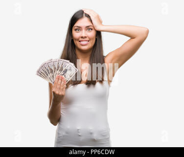 Young hispanic woman holding dollars a souligné avec la main sur la tête, choqué par la honte et la surprise face, en colère et frustrés. La peur et la colère pour mistak Banque D'Images