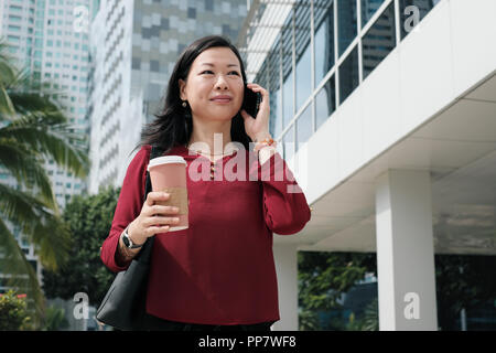 Happy Chinese female manager talking on cell phone et holding Coffee cup. Asian businesswoman walking au bureau le matin. Banque D'Images