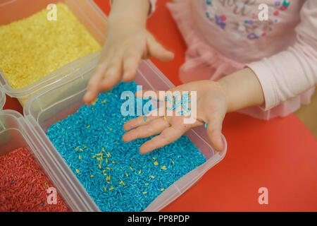 Les mains des tout-petits à jouer avec le riz arc-en-ciel dans la boîte sensorielle. Kit pédagogique sensoriel du bébé Banque D'Images