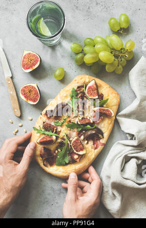 Pizza ou pain aux figues, roquette, prosciutto. Pizza d'automne avec des fruits et du jambon. Mains tenant la pizza sur fond de béton. Vue d'en haut, Banque D'Images