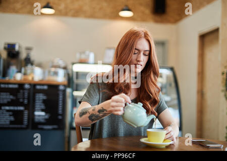 Red Head girl avec bras tatouage, assise seule dans un café se verse une tasse de thé comme elle s'assoit dans un café à Liverpool, Royaume-Uni Banque D'Images