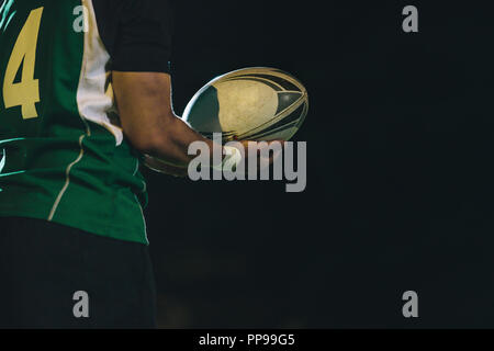 Plan recadré d'un joueur de rugby professionnel en uniforme tenant une balle dans un match de nuit. joueur de rugby avec ballon en main. Banque D'Images