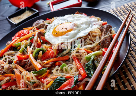 Un coréen sweet sour Japchae : Sauté de nouilles au boeuf, légumes et champignons shiitake, graines de sésame grillées, huile, sauce soja, dans une plaque noire Banque D'Images