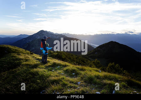Père et fils se tenant sur la montagne couverte d'herbe avec belle vue sur le coucher du soleil, en pointant sur le lac de Bohinj dans la vallée, Slatnik, Slovénie Banque D'Images