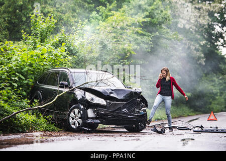Une jeune femme en colère avec le smartphone par la voiture endommagée après un accident de voiture. Banque D'Images