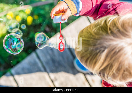 Vue aérienne de la partie supérieure de la tête d'un jeune enfant blond soufflant des bulles irisées colorées avec une solution de savon tout en se tenant à l'extérieur sur un bois Banque D'Images