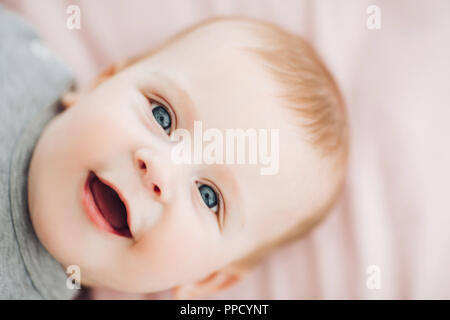 Close up of cute baby garçon aux grands yeux bleus, portant des vêtements en gris, le mensonge sur le lit et en regardant la caméra. Le repos, l'enfant de jouer. Concept de l'enfant et Banque D'Images