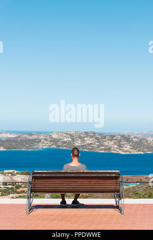 Young caucasian man, vu de dos, assis sur un banc à Palau, à la côte nord de la Sardaigne, l'Italie, l'observation de la mer Méditerranée et la M Banque D'Images
