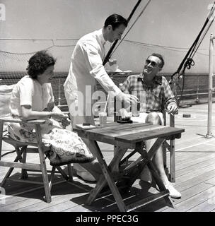 Années 1950, historique, un couple d'être servi un verre par un blanc adapté waiter tout en vous relaxant à l'extérieur, sur le pont d'un bateau à vapeur Union-Castle. Banque D'Images