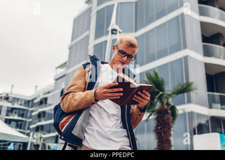 Sac à dos College student with reading book walking par cet hôtel moderne on tropical beach. Jeune homme étudiant à l'extérieur par temps pluvieux jour d'automne Banque D'Images