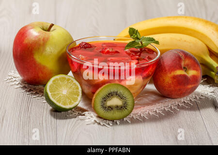 Gelée de cerises avec morceaux de fraises dans le bol en verre garni de feuilles de menthe, nectarine, kiwi, citron vert, pomme et banane sur la serviette blanche Banque D'Images