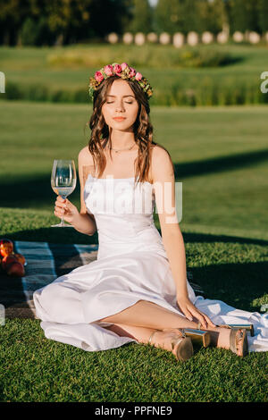 Belle Mariée offres en robe blanche et couronne holding glass of wine et assis sur couverture pour pique-nique romantique Banque D'Images